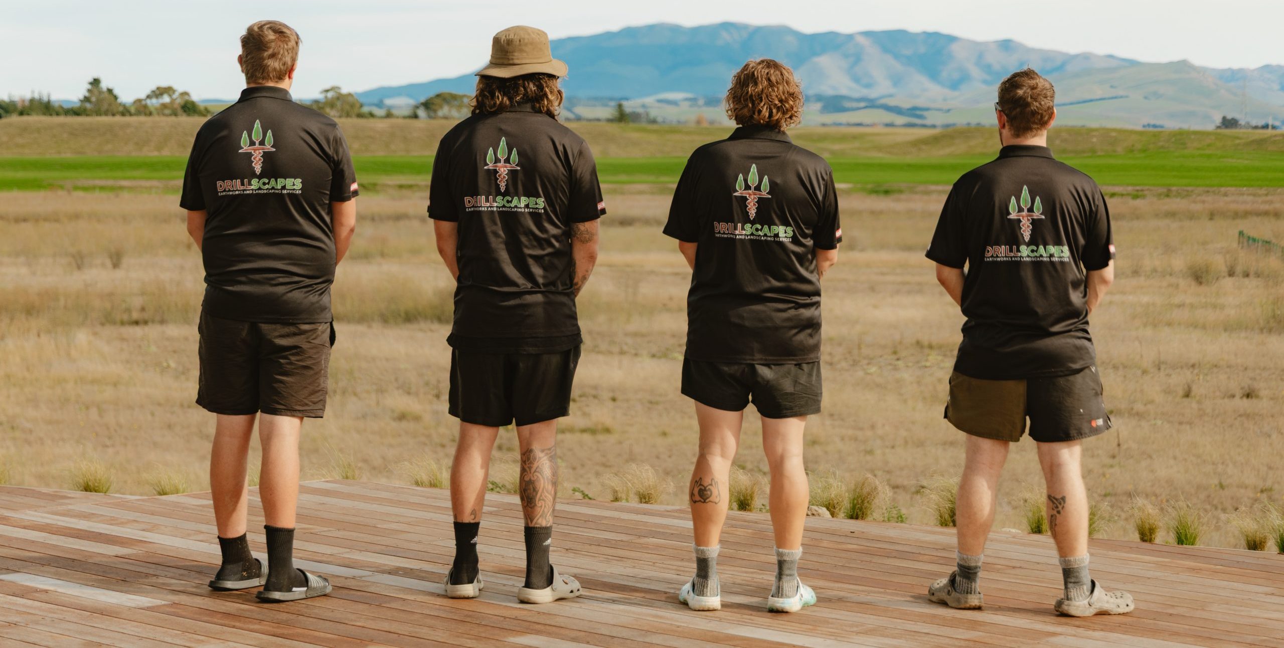 Drillscapes team standing on a wooden deck with their backs to the camera, wearing branded uniforms and looking out towards the North Canterbury mountains.”