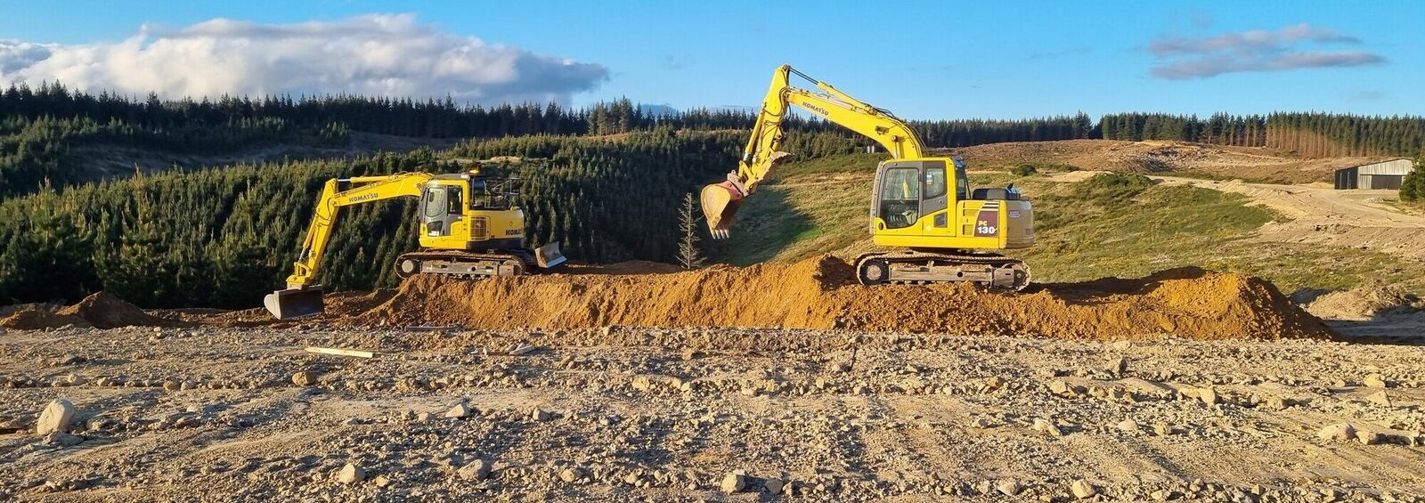 Two yellow Komatsu excavators working on a rural excavation site surrounded by pine forests and hills in North Canterbury under clear blue sky.
